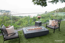 Load image into Gallery viewer, Elementi Hampton fire table lit outside on a patio with a windguard and tank enclosure
