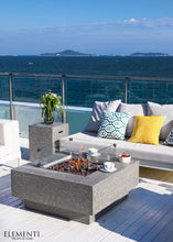 Load image into Gallery viewer, Elementi fire pit table on a beautiful deck with patio furniture and a flame and wind guard