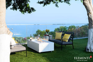 elementi carrara fire table with a flame and wind guard on a patio with a view of the ocean